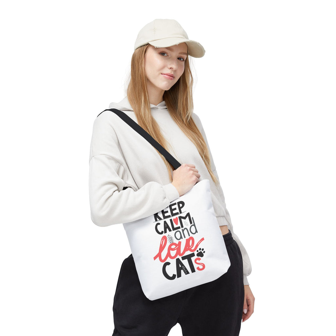 A woman wearing a beige cap, white hoodie, and black sweatpants carries a white canvas shoulder bag with the text 'KEEP CALM and love CATS' in black and red lettering, set against a plain white background.