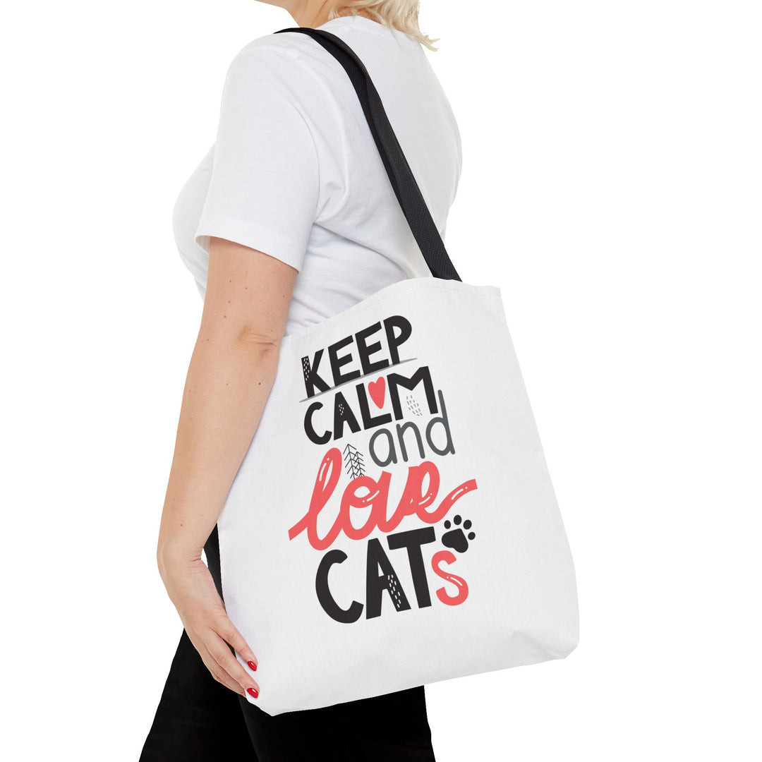 A white canvas tote bag with black and red text reading 'KEEP CALM and love CATS' is carried on the shoulder of a model wearing a white t-shirt and black pants, against a plain white background.