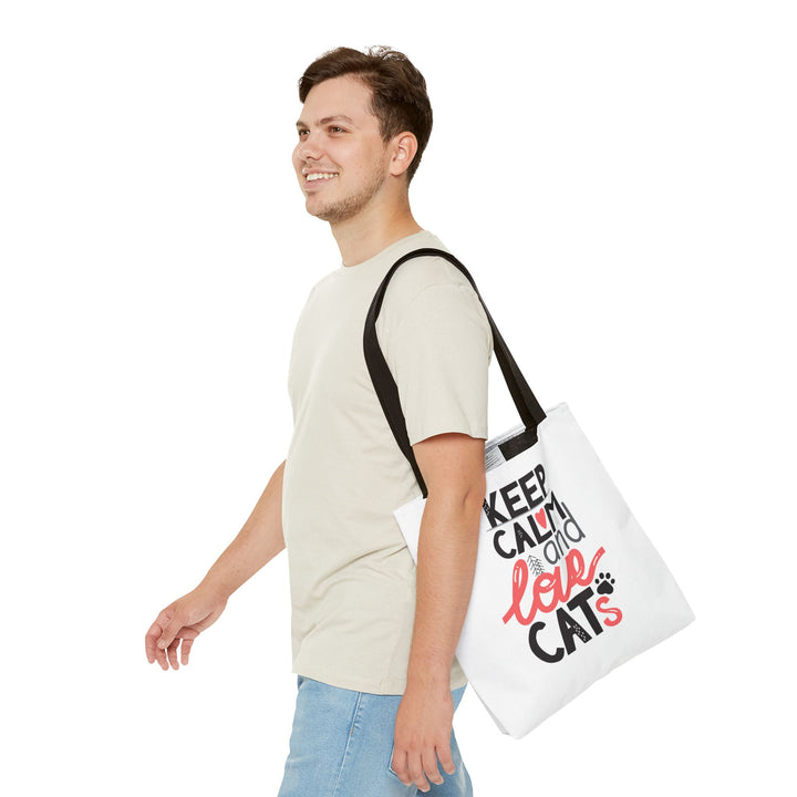 A man in a light beige t-shirt and blue jeans is shown from the waist up, carrying a white canvas tote bag over his shoulder. The bag features black straps and a graphic print that reads 'KEEP CALM AND LOVE CATS' in black and red lettering with a small black paw print.