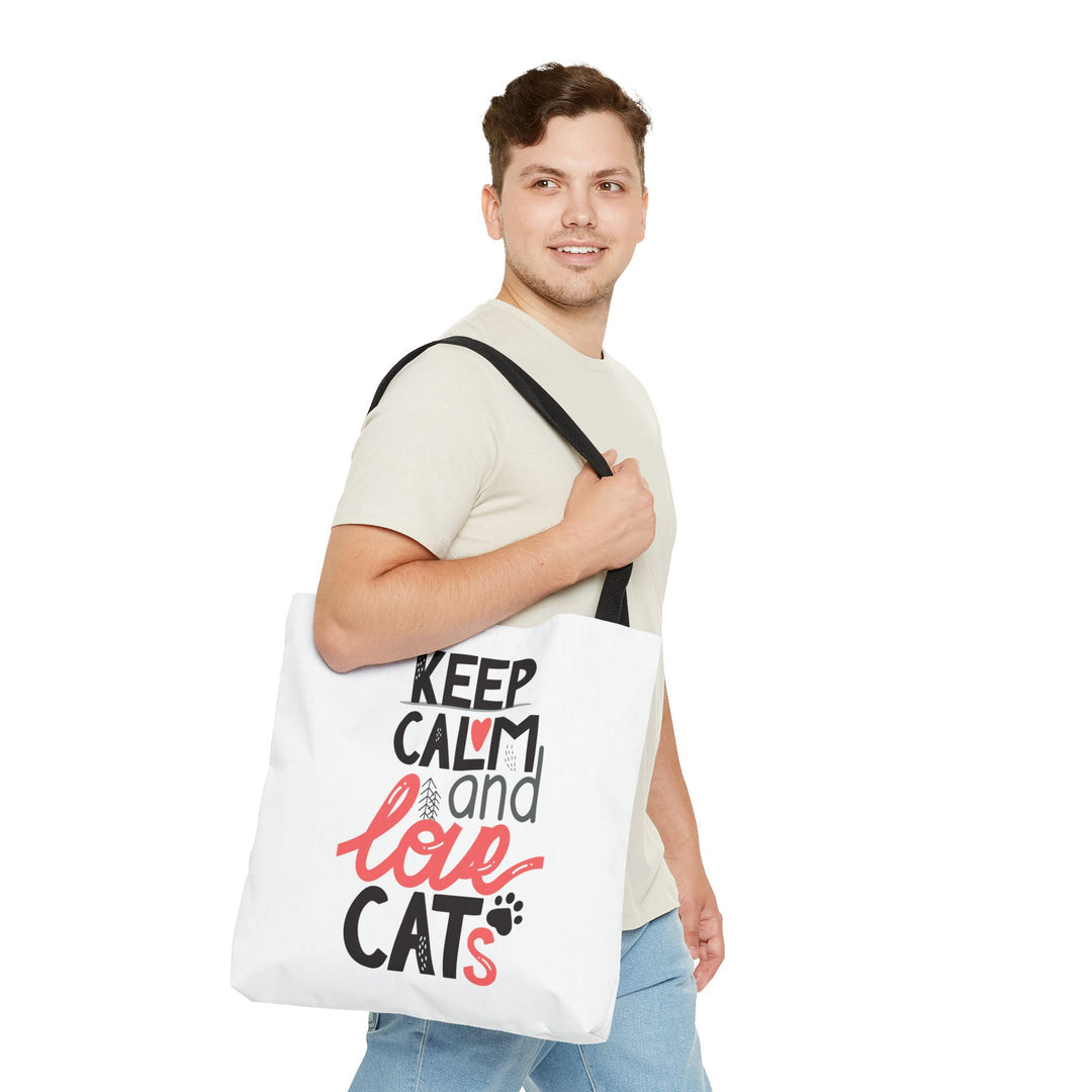 A man wearing a beige t-shirt and blue jeans carries a white canvas tote bag over his shoulder. The bag features the text 'KEEP CALM and love CATS' in black and red lettering with a small black paw print.