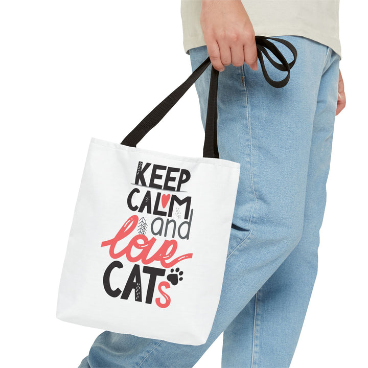 A white canvas tote bag with black handles, featuring the printed text 'KEEP CALM and love CATs' in black and coral pink, carried by a person wearing light blue jeans against a white background.