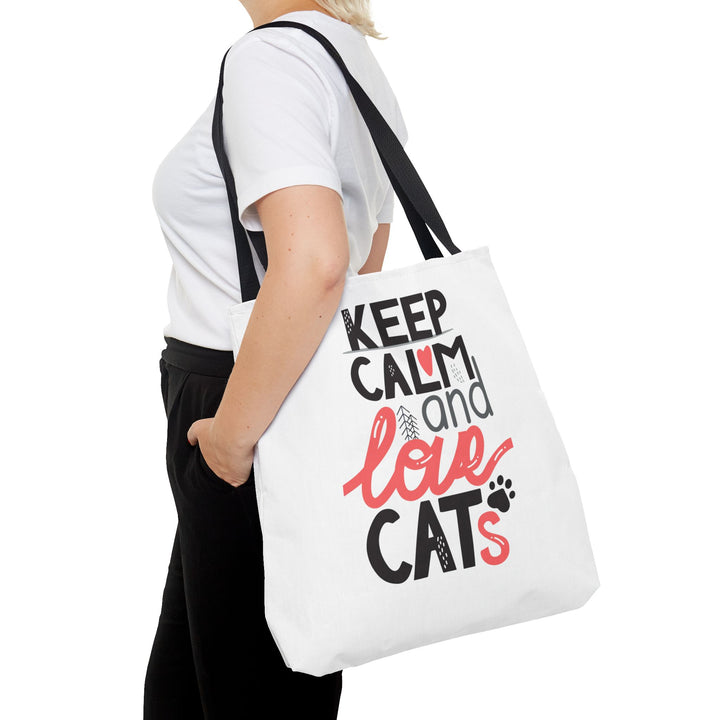 A white canvas tote bag with black and red text reading 'KEEP CALM and love CATS' with a small paw print, carried on the shoulder of a person wearing a white t-shirt and black pants against a white background.
