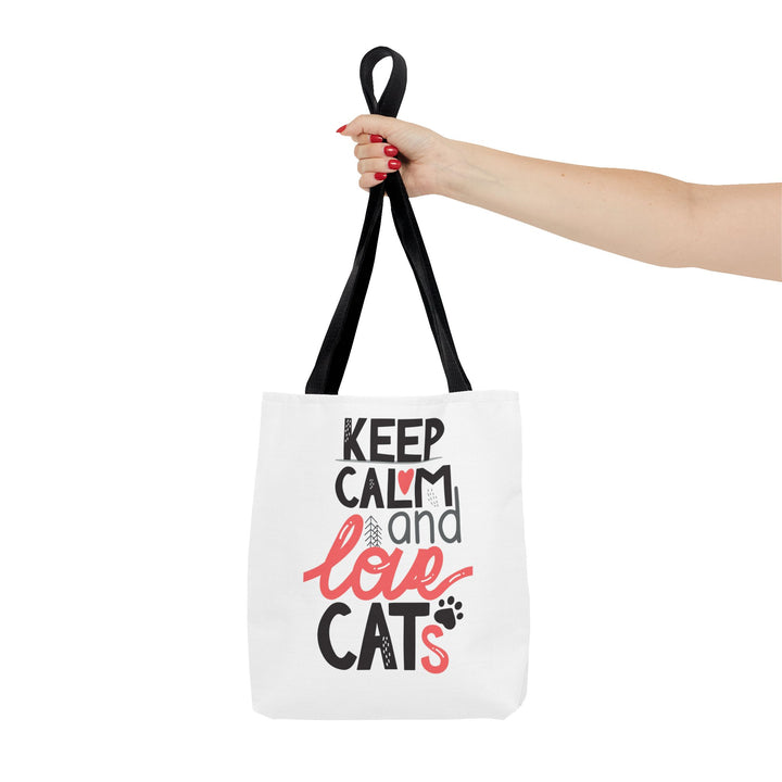 A white canvas tote bag with black handles, held by a person's hand against a plain white background. The bag features a graphic print that reads 'KEEP CALM and love CATS' in black and coral pink lettering.