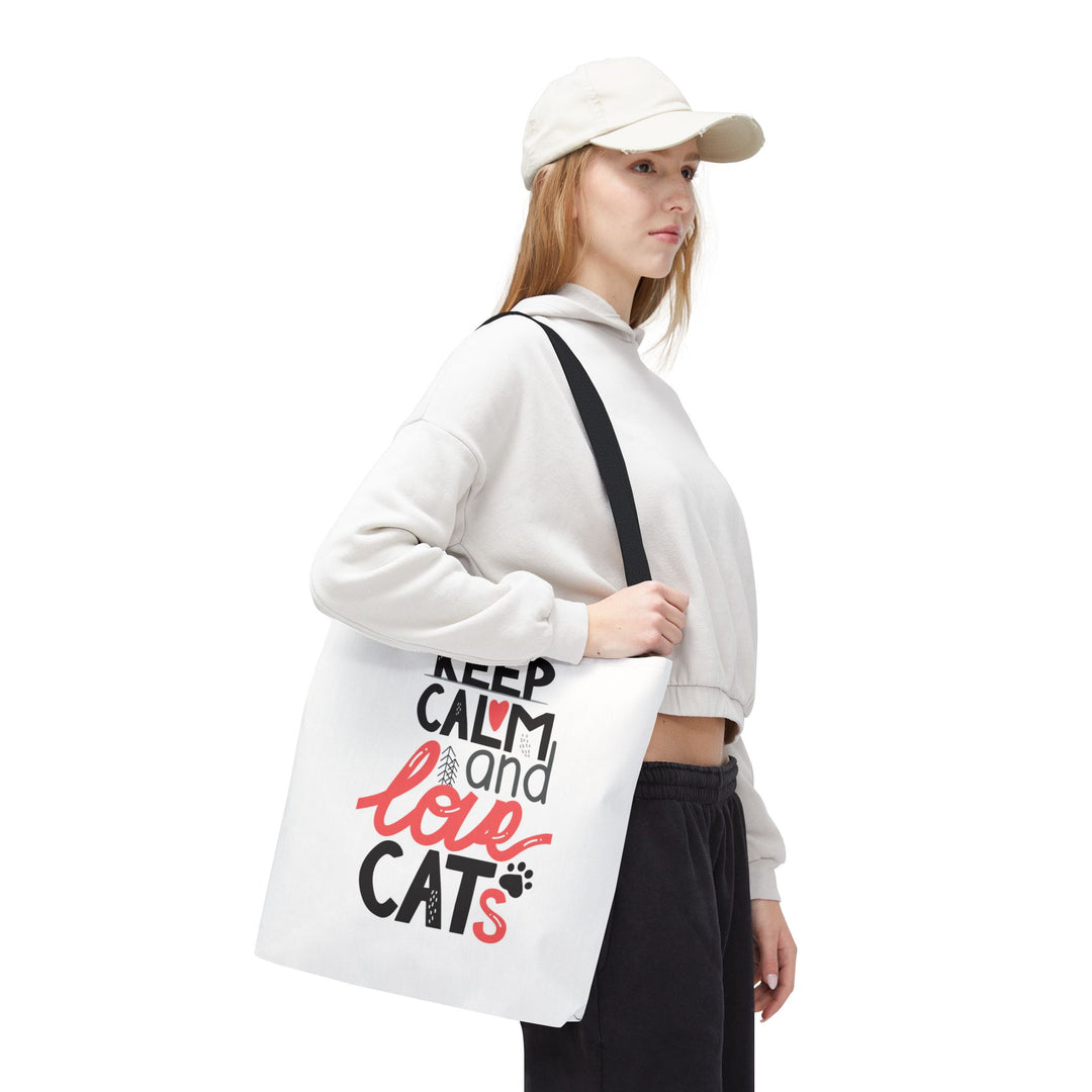 A woman wearing a white cap, white cropped hoodie, and black sweatpants carries a white canvas tote bag with a black shoulder strap. The bag features the printed text 'KEEP CALM and love CATS' in black and red lettering with a small cat silhouette and paw print.