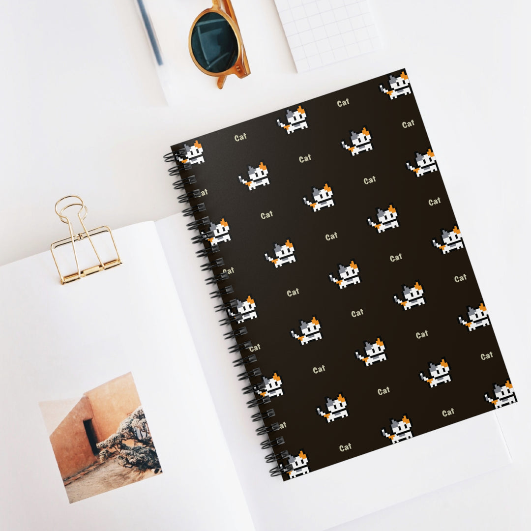 Video Game Cat Spiral Notebook displayed on a desk with sunglasses, a gold binder clip and photo, featuring pixel-art calico cats and repeating “Cat” lettering on the dark brown cover