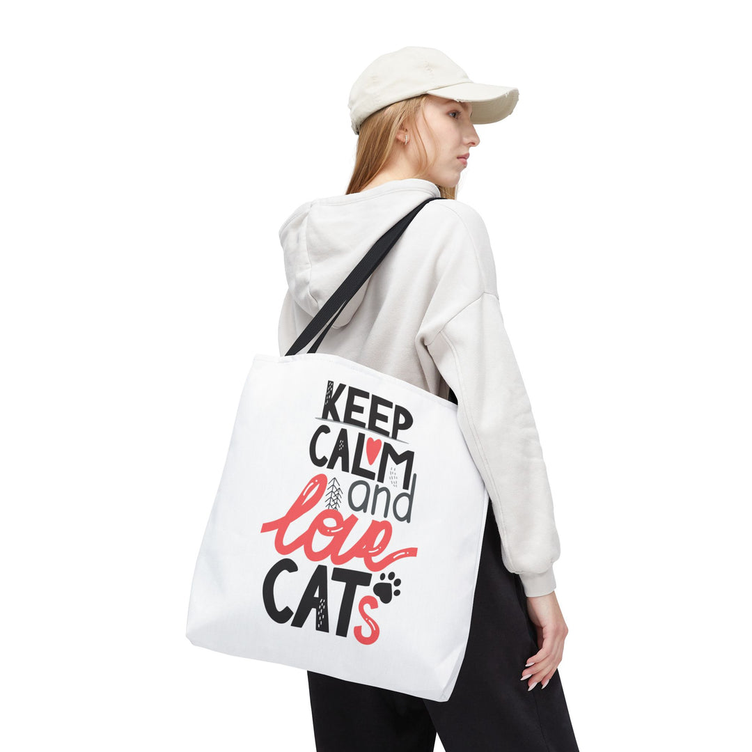 A woman wearing a white cap, white hoodie, and black pants is shown from the back while carrying a white canvas tote bag over her shoulder. The bag features the printed text 'KEEP CALM and love CATS' in black and red lettering with small cat and paw print details.