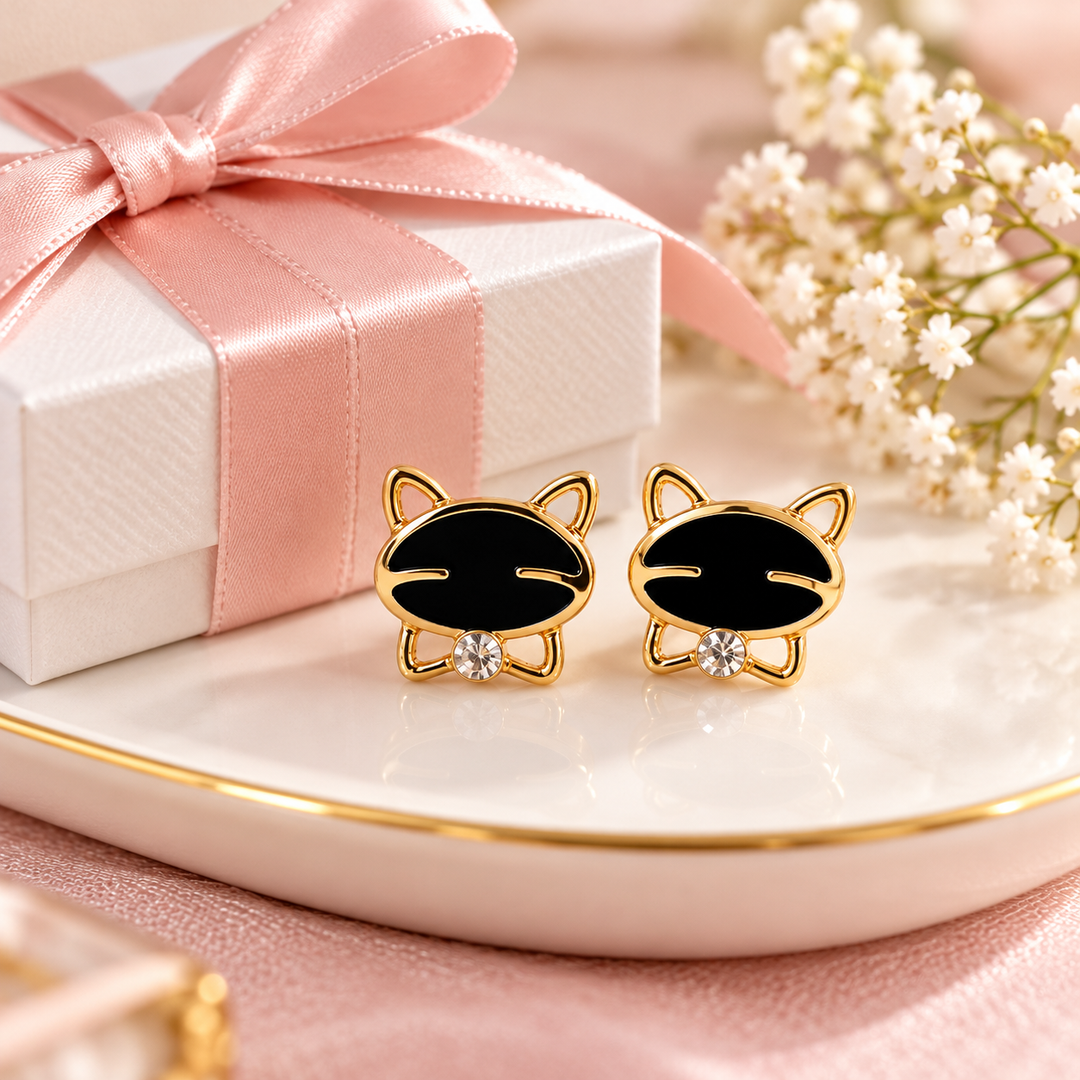 A pair of gold cat-shaped stud earrings with black enamel faces and a small clear crystal at the center, displayed on a white plate beside a pink gift box and white flowers.