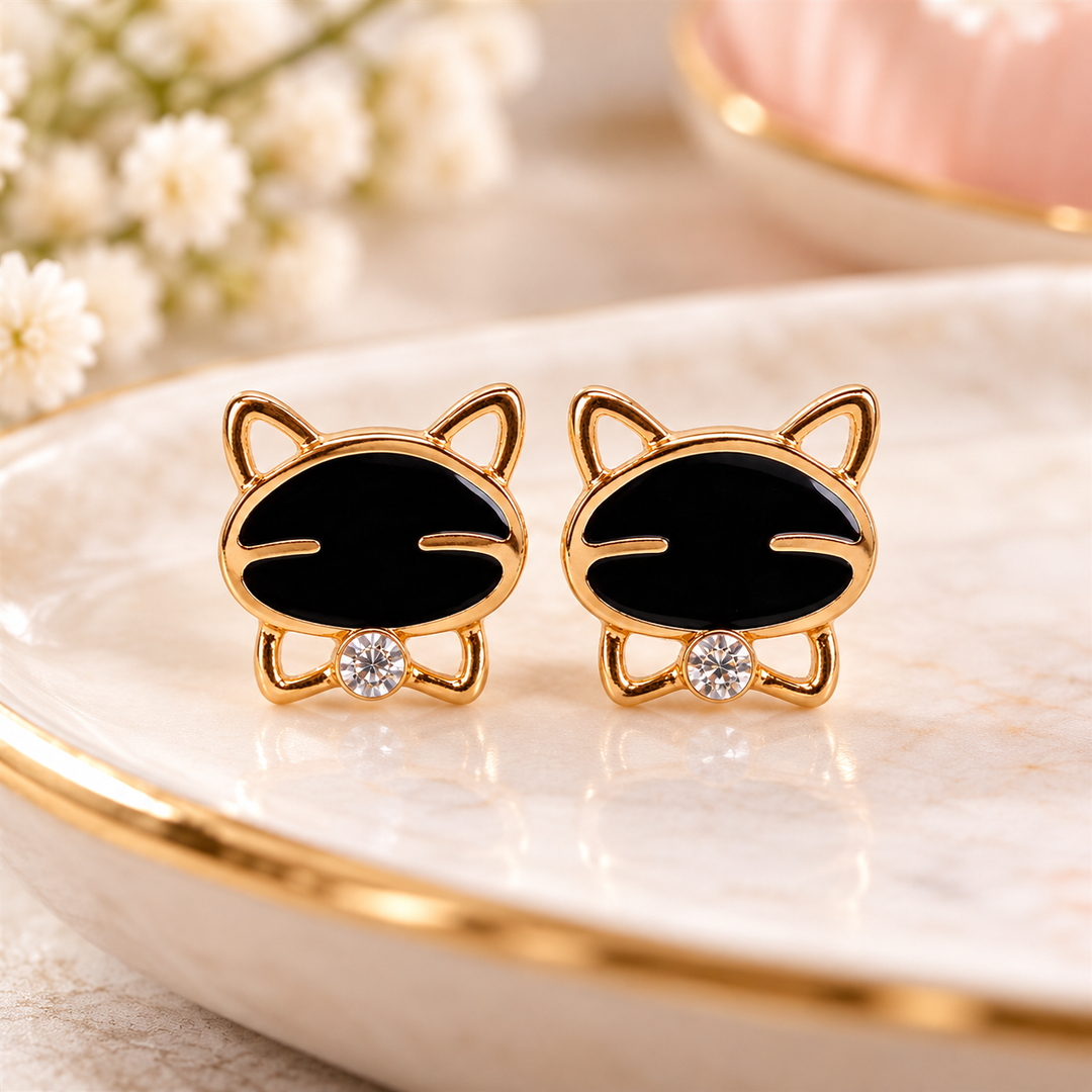 A pair of gold cat-shaped stud earrings with black enamel faces and small clear crystal accents, displayed on a white marble tray with gold trim.