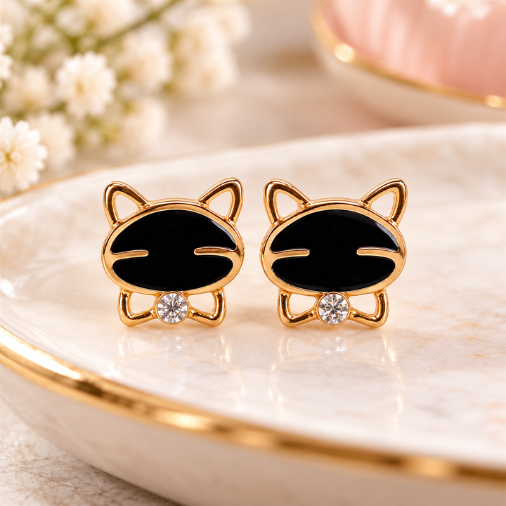 A pair of gold cat-shaped stud earrings with black enamel faces and small clear crystal accents, displayed on a white marble tray with gold trim.