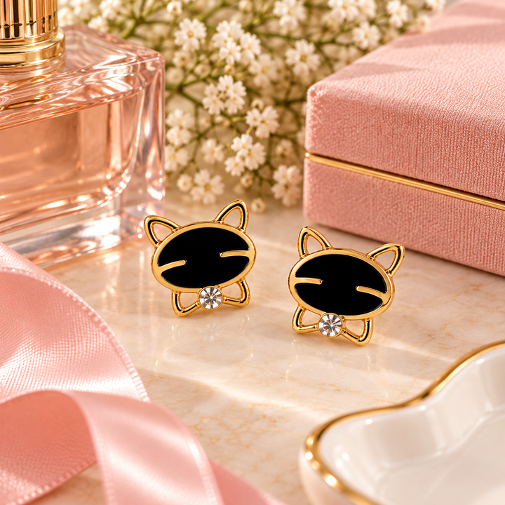A pair of gold-tone stud earrings shaped like cartoon cat heads with black enamel faces, displayed on a marble surface with a pink gift box, perfume bottle, flowers, and ribbon as styled props.
