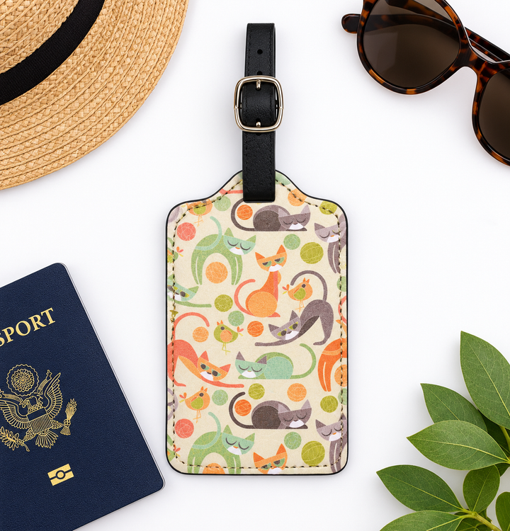 A rectangular luggage tag with a black strap and gold-tone buckle, featuring a colorful pattern of illustrated cats and citrus fruits on a cream background. The tag is styled in a flat lay with a navy passport, a straw hat, tortoiseshell sunglasses, and green leaves on a white surface.