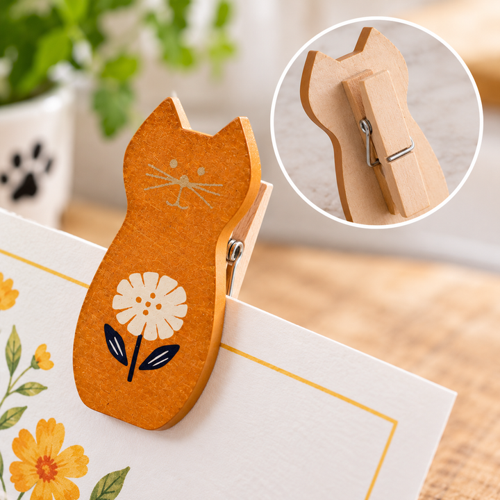 A wooden cat-shaped clothes pin with a white flower and black leaves design, holding a white card with yellow floral print. A circular inset shows the back of the pin with its metal spring and wooden body.