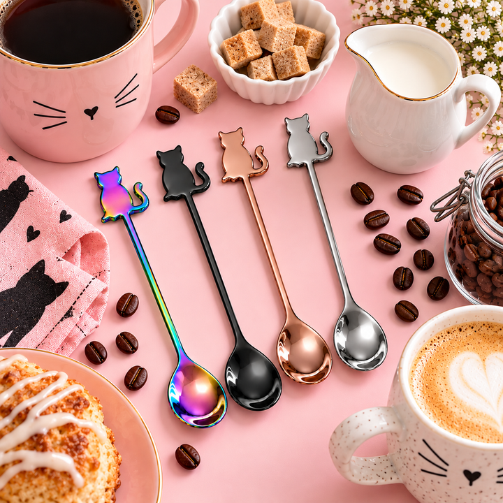 Four cat-shaped metal spoons in different finishes (iridescent rainbow, black, rose gold, silver) arranged on a pink background with coffee, pastries, and a milk pitcher.