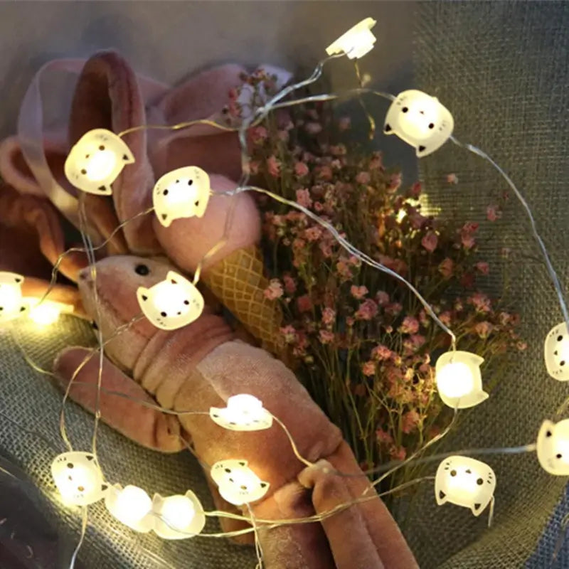 String lights shaped like cat faces draped over a bouquet of flowers and a plush toy.