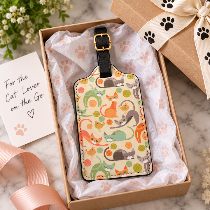 A rectangular cat-themed luggage tag with a black strap and gold buckle, displayed inside a brown gift box with tissue paper. The tag features a colorful pattern of illustrated cats in orange, green, gray, and black on a cream background.