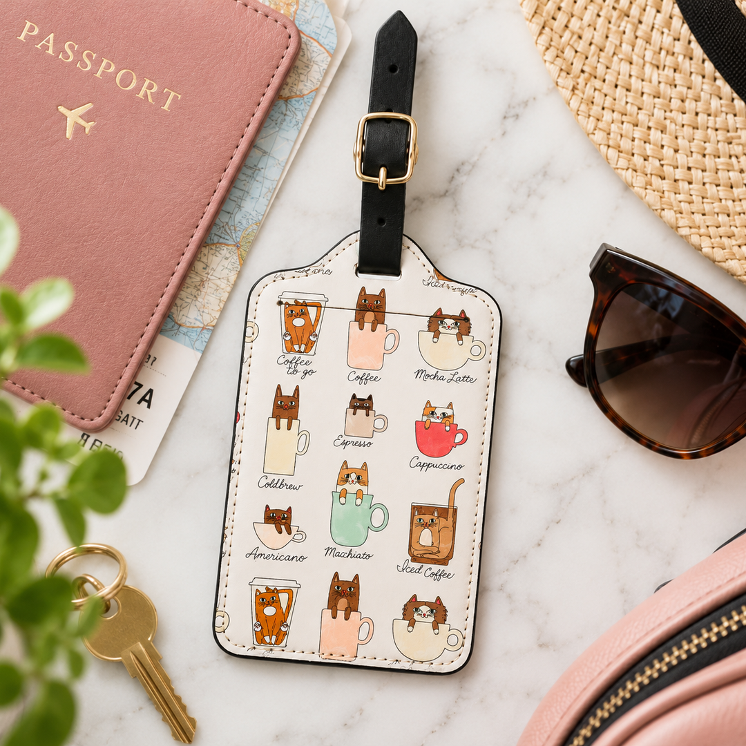 A rectangular luggage tag with a black strap and gold buckle, printed with a grid of illustrated coffee cups and cats, styled on a white marble surface with a pink passport, sunglasses, a straw hat, keys, and a pink bag nearby.