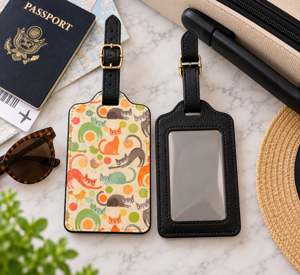 Two black faux-leather luggage tags displayed on a white marble surface. One tag features a colorful pattern of cartoon cats and circles on a cream background, while the other has a blank clear ID window.