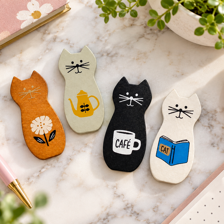 Four cat-shaped felt bookmarks or magnets arranged on a white marble surface, decorated with illustrations of a flower, a teapot, a coffee cup labeled 'CAFE', and an open book labeled 'CAT'.