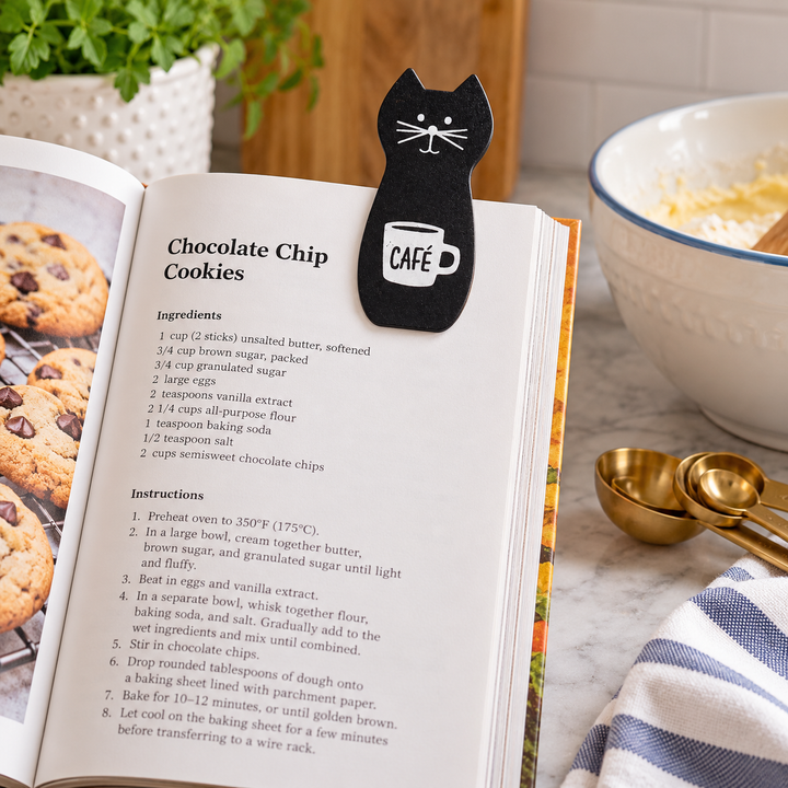 A black cat-shaped bookmark or clip with a white 'CAFE' mug graphic, holding open a cookbook titled 'Chocolate Chip Cookies' on a wooden table, with a bowl of cookies and gold spoons nearby.