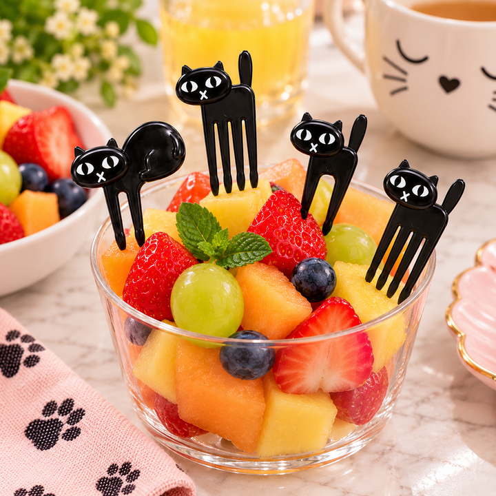 A clear glass bowl filled with mixed fruit, including strawberries, cantaloupe, grapes, and blueberries, decorated with four black cat-shaped plastic forks. The scene is set on a light marble surface with a pink paw-print cloth, a white cat-face mug, and other fruit bowls in the background.