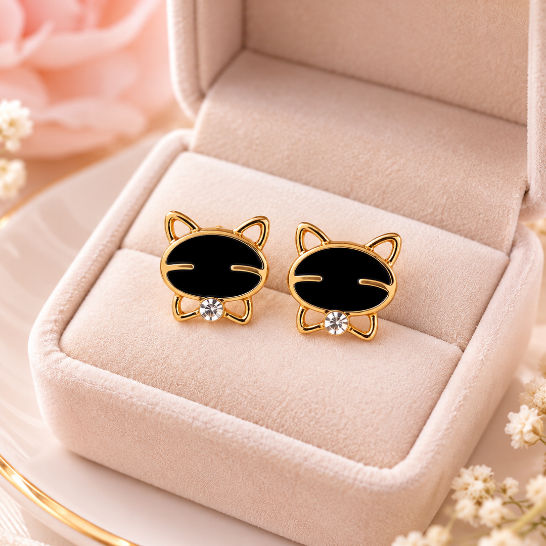A pair of gold cat-shaped stud earrings with black enamel faces and small clear crystal accents, displayed in a soft blush pink jewelry box.