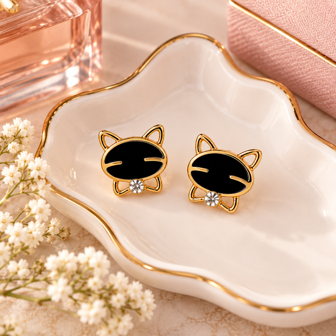 A pair of gold cat-ear stud earrings with black oval faces and small clear rhinestones, displayed on a white scalloped dish with gold trim.