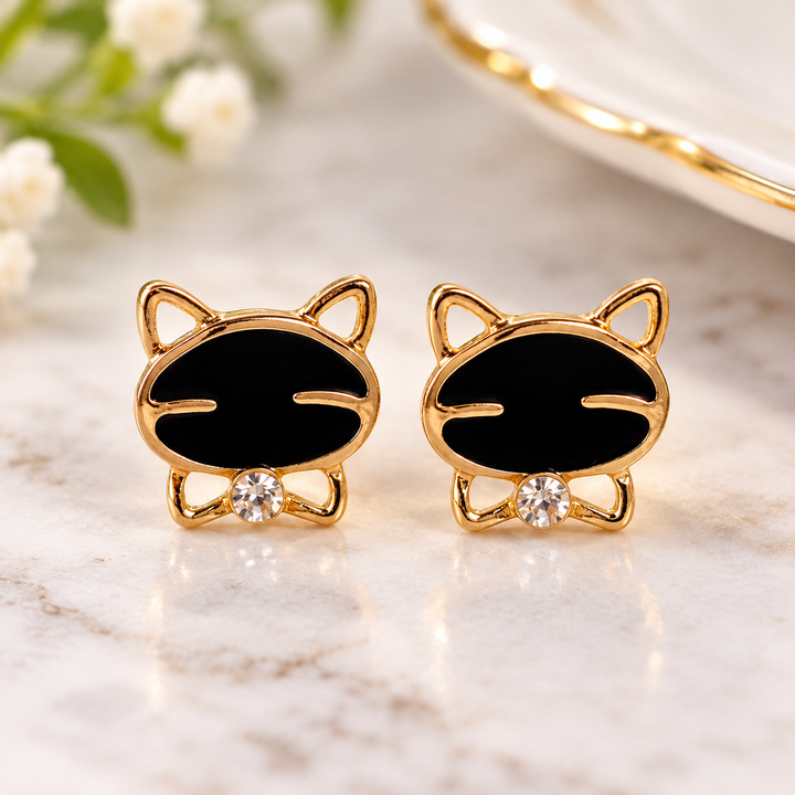 A pair of gold cat-shaped stud earrings with black enamel faces and small clear crystal accents, displayed on a light marble surface with soft floral background blur.