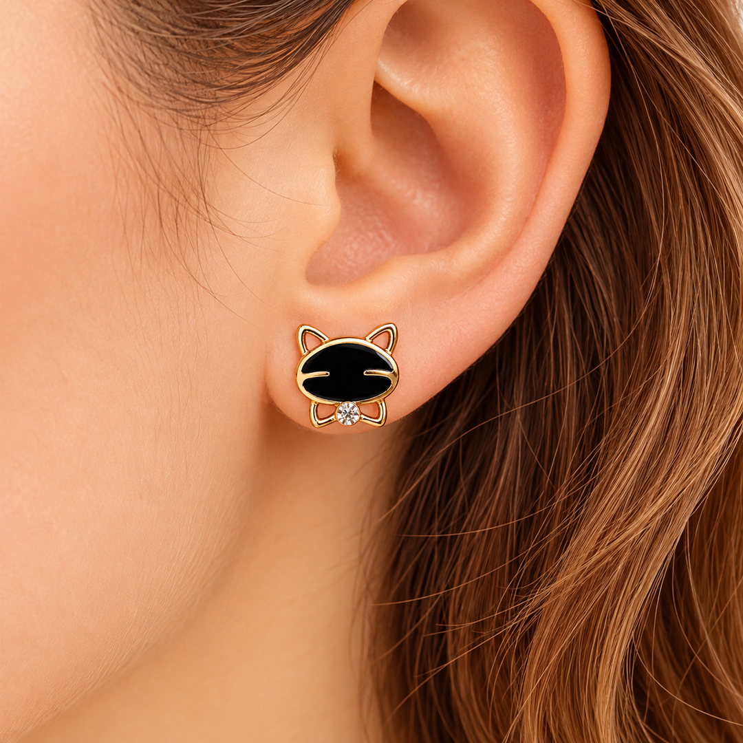 A close-up of a person's ear wearing a small cat-face stud earring with a black enamel face, gold outline, and tiny clear crystal detail.