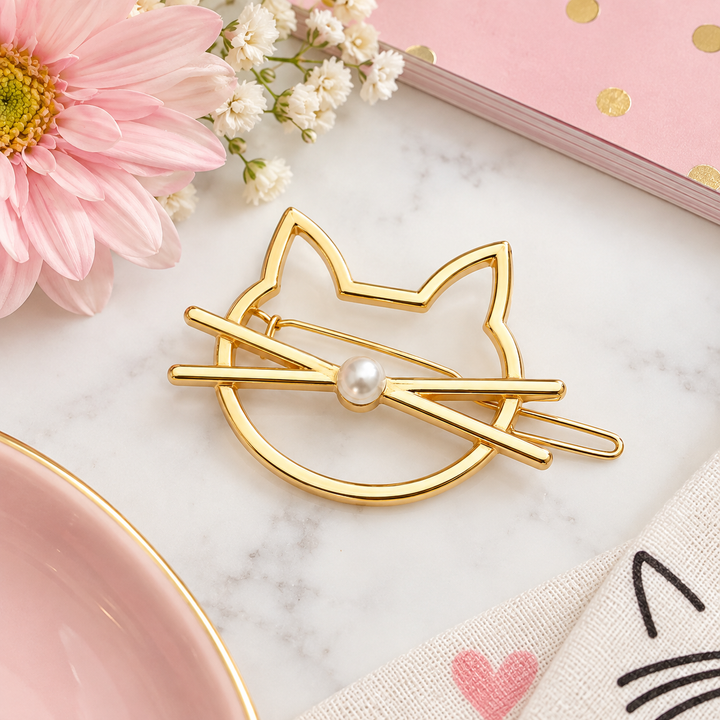 A gold cat-shaped hair claw clip with a white pearl in the center, styled on a white marble surface with a pink flower, white baby's breath, a pink notebook, and a beige fabric napkin.