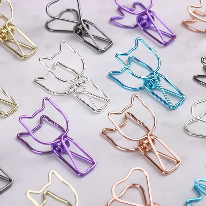 A close-up of several metal paperclips arranged on a light marble-like surface. The clips are made of thin wire in multiple finishes, including gold, silver, purple, blue, and rose gold.