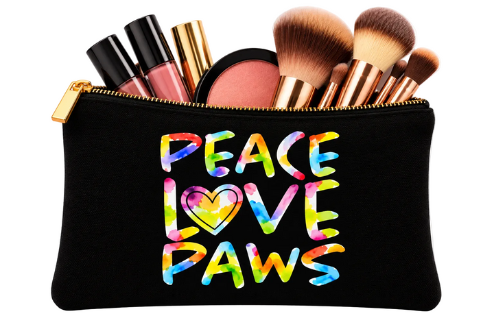 A black cosmetic pouch with a gold zipper, filled with makeup brushes, lip products, and a compact. The front of the pouch features the text 'PEACE LOVE PAWS' in a colorful tie-dye style with a heart in the center, set against a plain white background.