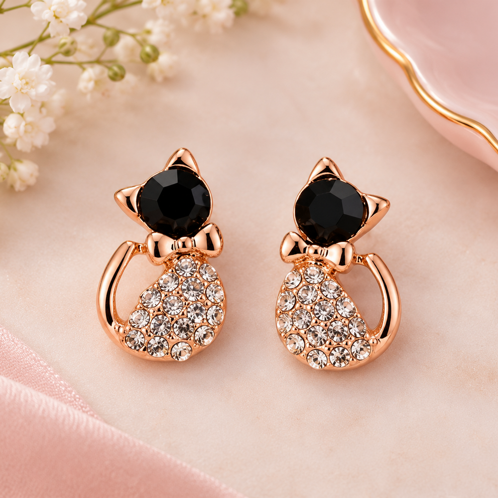 A pair of rose gold cat-shaped stud earrings with black faceted gemstones on the heads and crystal-encrusted bodies, displayed on a soft pink satin surface with baby's breath flowers and a pink dish in the background.