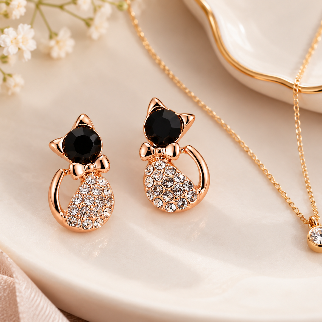 A jewelry set featuring a pair of rose gold cat-shaped stud earrings with black faceted gemstones and crystal-encrusted bodies, displayed alongside a matching gold chain necklace with a round crystal pendant on a white ceramic dish with baby's breath flowers.