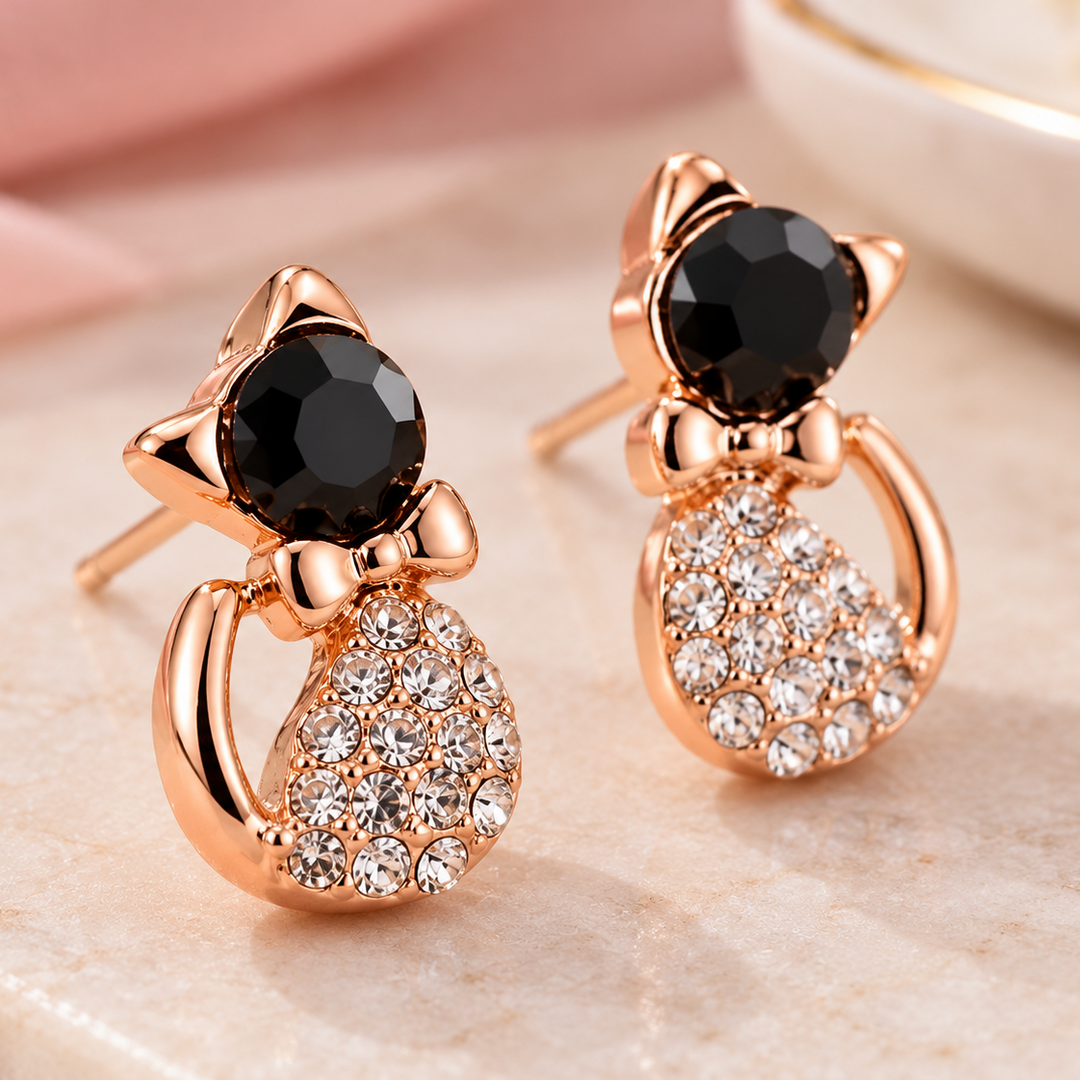 A pair of rose gold cat-shaped stud earrings with black faceted gemstones and clear rhinestone detailing, displayed on a light marble surface with soft pink fabric in the background.