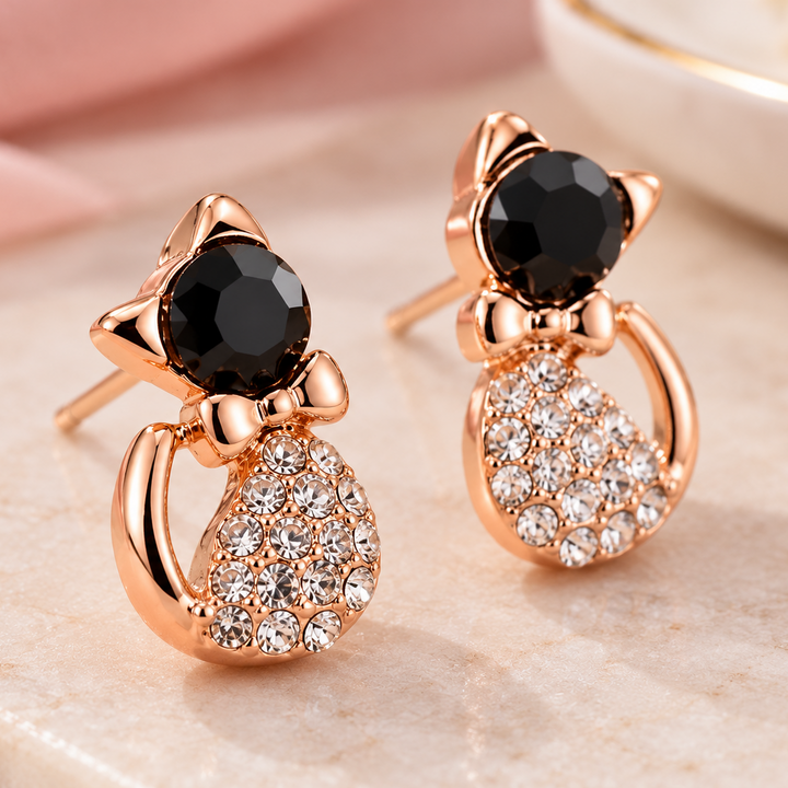 A pair of rose gold cat-shaped stud earrings with black faceted gemstones and clear rhinestone detailing, displayed on a light marble surface with soft pink fabric in the background.