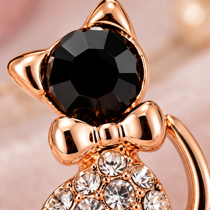A close-up of a rose gold cat-shaped ring featuring a large faceted black gemstone on the forehead, a bow detail, and clear rhinestones on the body, set against a soft pink background.