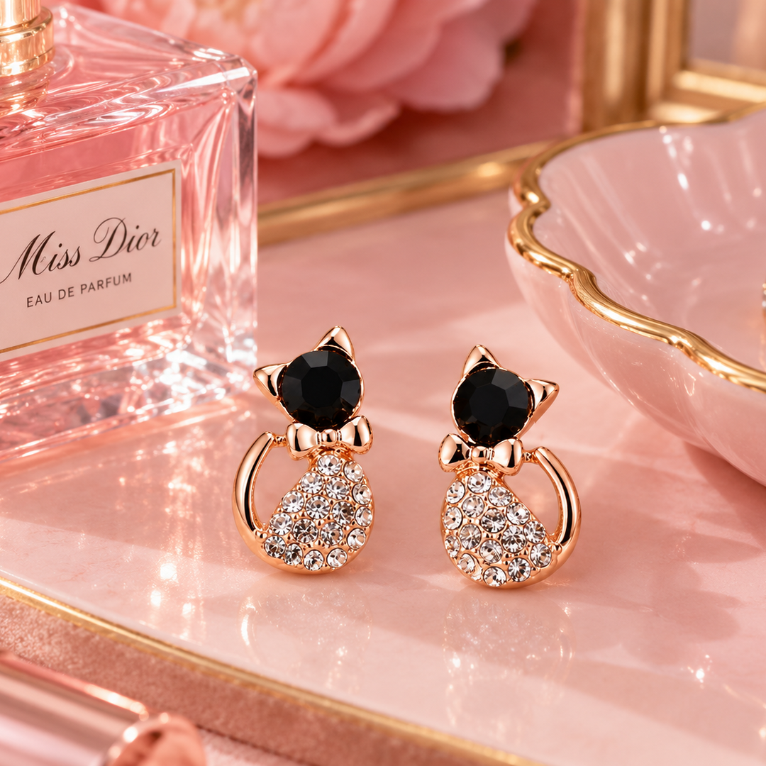 A pair of rose gold cat-shaped stud earrings with black faceted gemstones on the heads and clear crystal-encrusted bodies, displayed on a pink tray next to a Miss Dior perfume bottle.