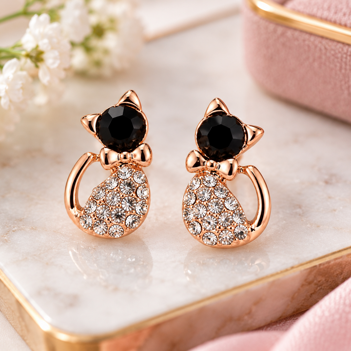 A pair of rose gold cat-shaped stud earrings with black faceted gemstones on the heads and crystal-encrusted bodies, displayed on a marble tray with white flowers and a pink fabric background.