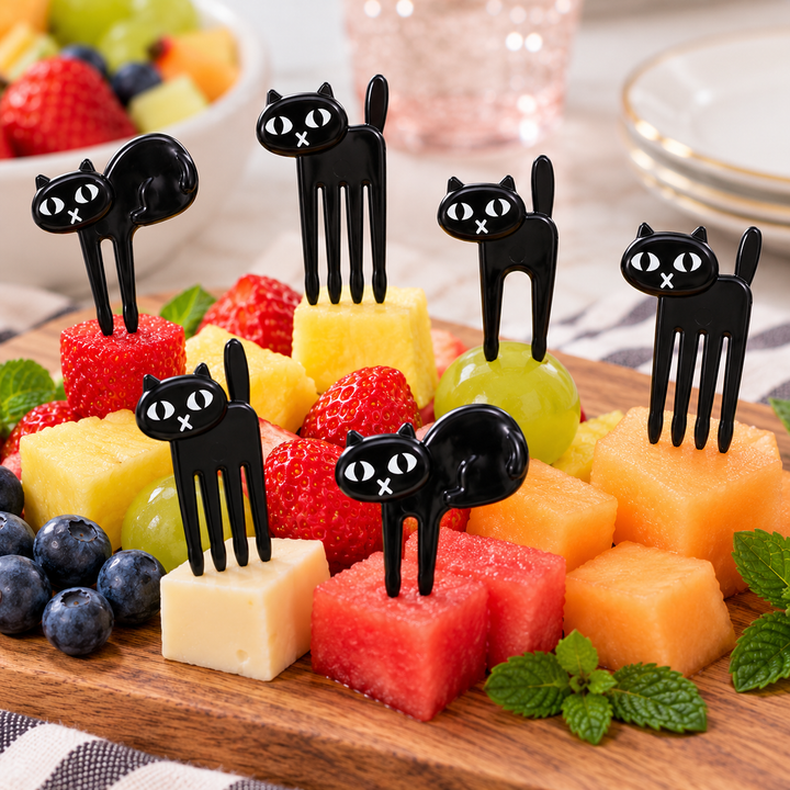Five black cat-shaped plastic fork picks with white eyes are displayed on top of colorful fruit and cheese cubes on a wooden board.
